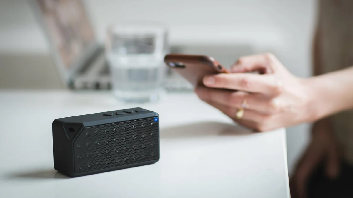 Mão segurando smartphone próximo a uma caixinha de som Bluetooth portátil em cima da mesa
